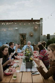 a group of people sitting around a wooden table eating food and drinking wine at sunset