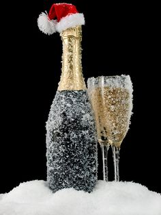 two champagne flutes and a bottle of champagne in the snow with a santa hat on top