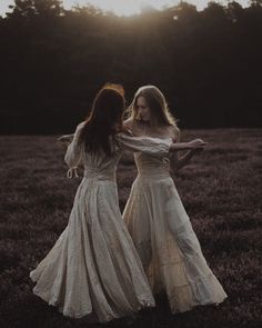 two women in long dresses are standing in a field at sunset or sunrise, with the sun shining behind them