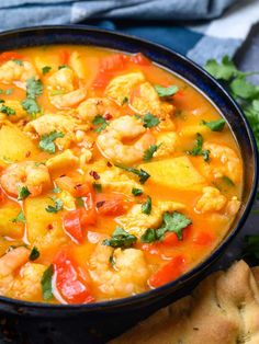 a bowl filled with shrimp and vegetables next to bread on a blue table cloth, surrounded by cilantro