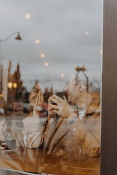 two men sitting at a table in front of a window talking on their cell phones