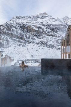 a person in a large hot tub with snow on the mountain behind it and buildings