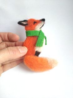 a hand is holding a small felt fox with a green scarf around it's neck