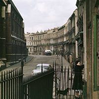 a woman standing on the side of a road next to a fence and cars parked in front of buildings