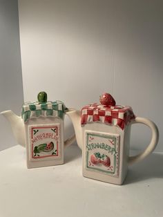 two ceramic teapots sitting next to each other on a white counter top with a checkered cover