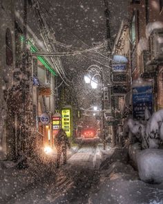 a person walking down a snowy street at night with snow falling on the ground and buildings in the background