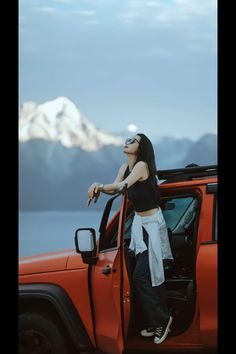 a woman standing in the open door of a red jeep with mountains in the background