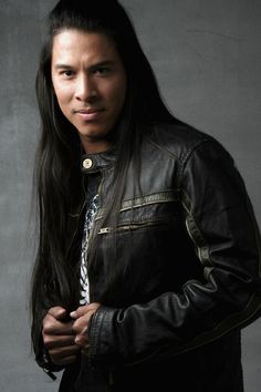 a man with long black hair wearing a leather jacket and standing in front of a gray background