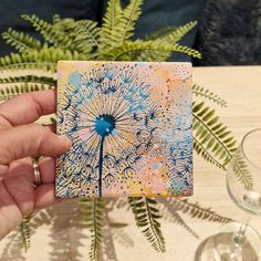 a person holding up a small card with a dandelion design on it, sitting at a table