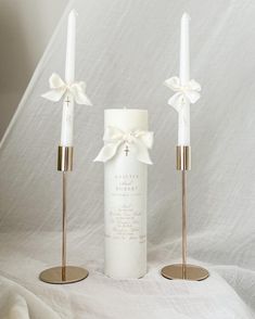 two white candles with bows on them sitting next to each other in front of a white sheet