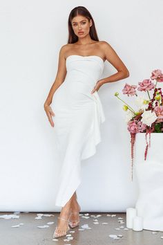 a woman in a white dress standing next to a vase with flowers on the floor