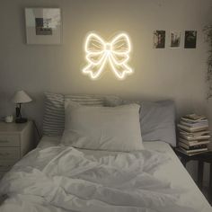 a white bed topped with pillows under a neon bow wall light next to a night stand