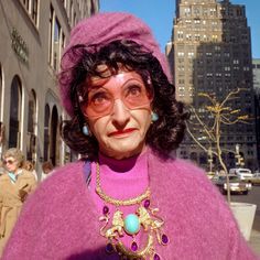 an old woman wearing glasses and a purple sweater in front of tall buildings with people walking down the street