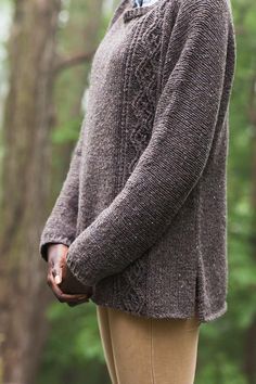 a woman standing in the woods with her hands on her hips wearing a brown sweater and tan pants