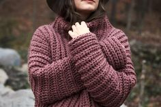 a woman wearing a hat and sweater standing in front of rocks with her hands on her chest