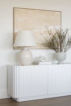 a white table with a lamp and vase on it in front of a large painting