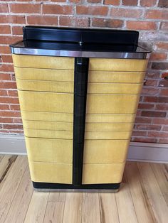 a yellow and black trash can sitting on top of a hard wood floor next to a brick wall