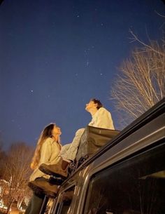 two people sitting on the roof of a car looking up at the stars in the sky