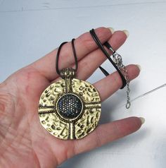 "This fabulous and eye-catching 1980s vintage black neck cord necklace has a large 2\" wide round hammered antiqued gold-tone pendant with a gorgeous Etruscan Revival design.  It's center has amazing black and blue aurora borealis rhinestones that have brilliant gold sparkle.  The black neck cord is 18\" long plus it has a 2\" extender chain.  The back of the pendant looks like it has wear, but it feels smooth and solid so I think it was made that way to look antique.  It is in excellent conditi Black Metal Necklace With Antique Finish, Black Metal Jewelry With Antique Finish, Black Metal Medallion Necklace, Black Metal Medallion Jewelry, Black Medallion Metal Jewelry, Adjustable Metal Medallion Necklace, Round Metal Medallion Necklace With Large Pendant, Revival Design, Blue Aurora