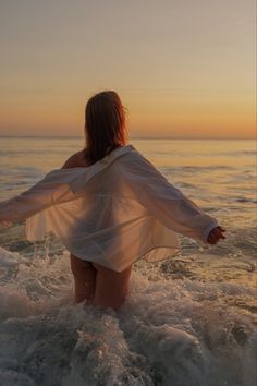 a woman standing in the ocean at sunset with her arms spread out and back to the camera