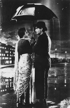 a man and woman standing under an umbrella in the rain with buildings in the background