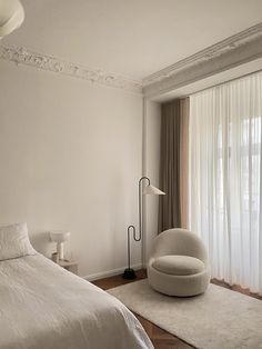 a bedroom with a white bed, chair and large window in the corner that has sheer curtains on both sides