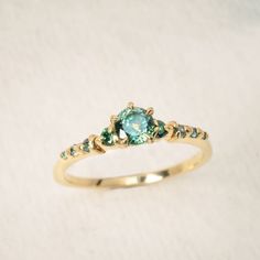 a gold ring with an aqua blue stone and green stones on the side, sitting on a white surface