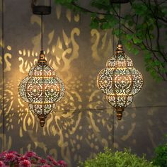 two decorative lights hanging from the side of a wall next to flowers and trees in front of a building