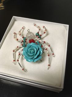 a white box with a blue rose and beaded spider on it's side