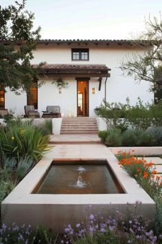 an outdoor fountain in front of a house with flowers and plants around it, surrounded by greenery