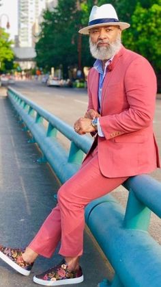 an older man in a pink suit and hat sitting on a blue metal rail with his legs crossed