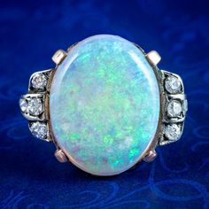 A striking antique Victorian opal statement ring boasting a magnificent natural opal cabochon weighing approx. 8 carats. It exhibits a changing kaleidoscope of greens, blues and purples with a warm orange glow behind. It's flanked by blocked shoulder accents dotted with six twinkling diamonds and further diamonds chasing into the band (approx. 0.30ct total).  Opal is the birthstone of October. Their magic has been revered throughout history, desired for their enchanting rainbow of colours that f Antique Oval Opal Gemstone Ring, Antique Oval Opal Ring, Heirloom Opal Ring Collectible, Heirloom Style Collectible Opal Ring, Antique Oval Multi-stone Opal Ring, Antique Style Opal Ring For Formal Occasions, Victorian Style Opal Ring For Formal Occasions, Heirloom Opal Cabochon Ring Collectible, Antique Style Round Opal Ring