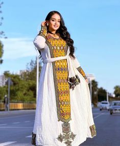 a woman in a white and yellow dress standing on the side of a road with her hands behind her head