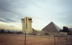 a tall building sitting next to a large pyramid on top of a dirt field in front of a fence