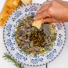 a person is dipping some bread into a bowl of olives and herbs on a blue and white plate