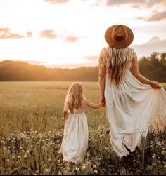 a mother and daughter holding hands walking through a field with the sun setting in the background