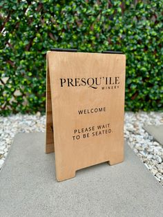 a wooden sign that says, welcome please wait to be seated in front of some bushes