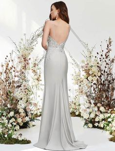 the back of a woman in a long silver dress standing next to flowers and trees