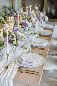 a long table is set with white plates and purple flowers on it, along with candles