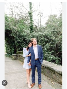 a man and woman standing next to each other in front of some green trees on the sidewalk