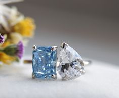 two engagement rings with blue and white diamonds on them, next to some wildflowers