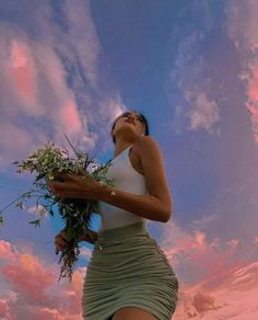 a woman in a white top and green skirt is holding some flowers while looking up into the sky