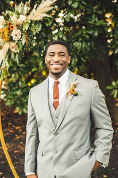 a man in a gray suit and tie standing next to a flower arch with an orange boutonniere on it