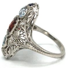 This fascinating Edwardian ring—circa the 1910s—is made of platinum and features four different stones of four different colors. Crafted in the elongated or "navette" style, a central checkerboard topped red garnet is supported by a pair of horizontally oriented antique oval cut aquamarines and six single cut diamonds. Punctuating both the top and bottom of the ring is a pair of deep-blue chunky round cut sapphires. The space between the stones is filled with the lacy filigree so beloved of the Antique Oval Multi-stone Ring, Art Deco Multi-stone Oval Jewelry, Classic Multi-stone Marquise Rings, Classic Marquise Multi-stone Ring, Antique Oval Three Stone Rings, Art Deco Platinum Multi-stone Rings, Art Deco Multi-stone Platinum Rings, Platinum Multi-stone Art Deco Rings, Heirloom Three Stone Marquise Ring