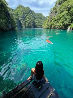 a woman is sitting on a bench looking out at the blue water and people swimming in it