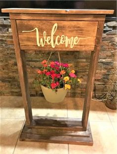 a welcome sign with flowers in a pot on a stand next to a stone wall