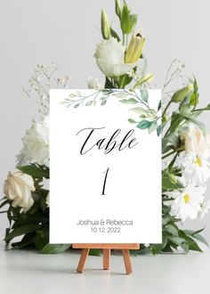 a table number card with white flowers and greenery in the background on an easel