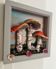 two mushrooms are in a shadow box with flowers on the bottom and one mushroom is painted red