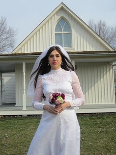 a woman in a white wedding dress standing on the grass outside of a church wearing a veil and holding a bridal bouquet