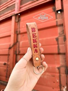 a hand holding a keychain with the word beka on it in front of a red wall